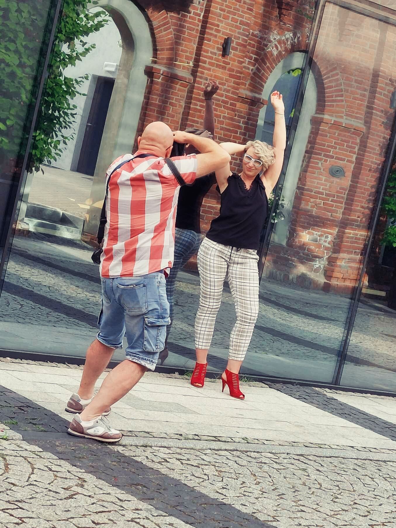 Fotograf w akcji podczas sesji w miejskiej scenerii. Mężczyzna w koszuli w czerwoną kratę i jeansowych szortach fotografuje roześmianą kobietę w ciemnym topie i wzorzystych spodniach, która pozuje z podniesionymi rękami na tle ceglanego budynku z dużymi, przeszklonymi witrynami.