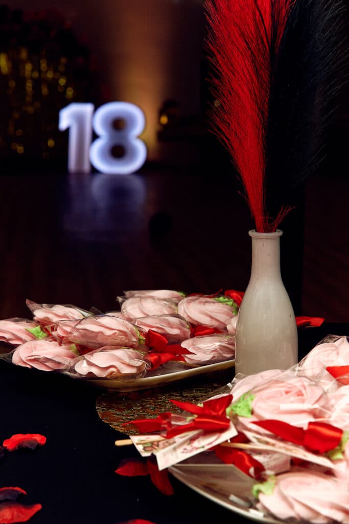 Dekoracje z motywem '18' na stole urodzinowym. Na pierwszym planie widoczne są talerze z bezami w kształcie róż, owiniętymi w folię i przewiązanymi czerwonymi wstążkami, oraz biały wazon z czerwonymi piórami. W tle, na ciemnej powierzchni, stoi podświetlana biała cyfra '18', tworząc centralny element dekoracji.