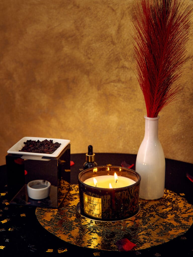 Dekoracyjny świecznik i aromaty na 18. urodzinach. Na stole pokrytym czarnym obrusem ze złotym wzorem stoi zapalona, wieloświecowa świeca w ciemnym szkle. Obok znajduje się ceramiczny aromatyzer z ciemnymi granulkami oraz biały wazon z czerwonymi piórami. W tle widać ścianę o złotej, teksturowanej powierzchni.