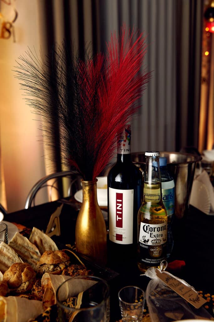 Dekoracje stołu na 18. urodzinach. Na czarnym obrusie stoi złoty wazon z czarno-czerwonymi piórami. Obok znajdują się butelki wina Martini i piwa Corona Extra, a także metalowy kubełek na lód. Na pierwszym planie widoczne są talerze z przekąskami. W tle widać fragment zasłon i oświetlenia.