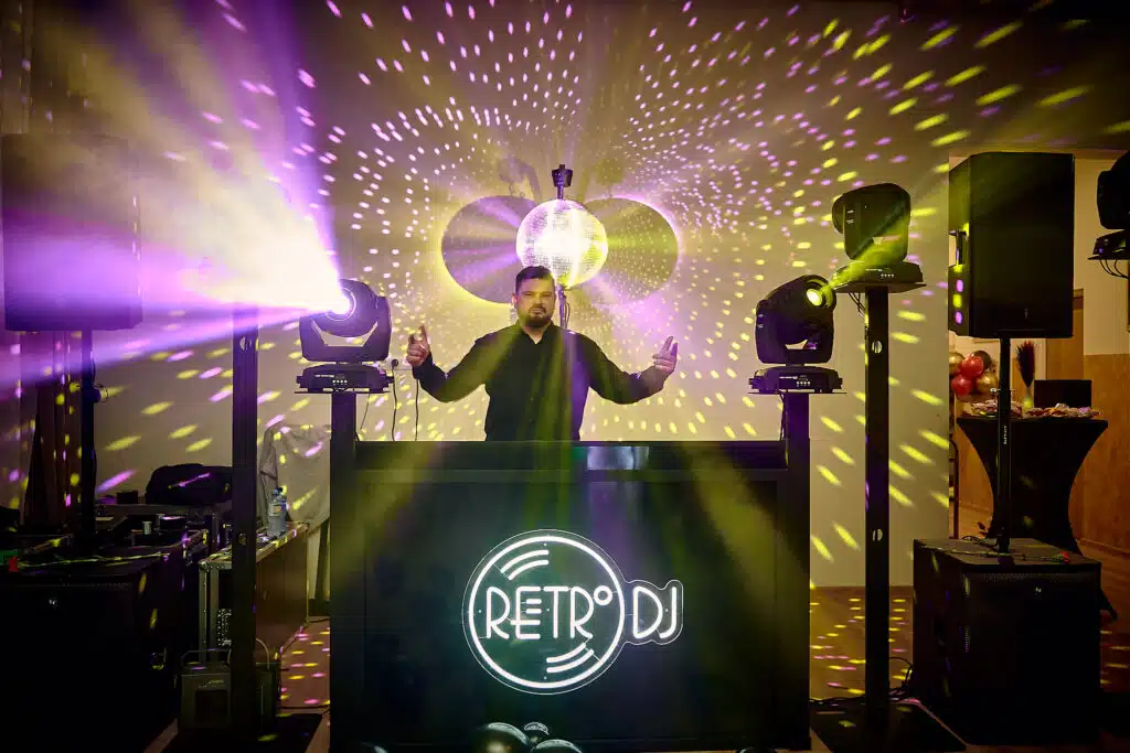 DJ na 18. urodzinach w akcji. Mężczyzna stoi za konsoletą z dużym logo 'RETRO DJ', z uniesionymi rękami, w otoczeniu profesjonalnego oświetlenia dyskotekowego. Wokół niego widać reflektory, kolumnę głośnikową oraz kule dyskotekowe rzucające wzory na ścianę i sufit.