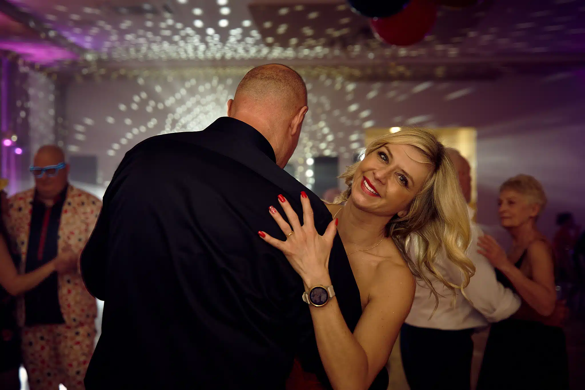 Rodzice jubilata tańczą podczas 18. urodzinowej imprezy. Kobieta z blond włosami uśmiecha się, trzymając rękę na ramieniu mężczyzny w czarnej koszuli, który stoi do niej tyłem. W tle widać bawiących się gości i odbijające się światła dyskotekowe.
