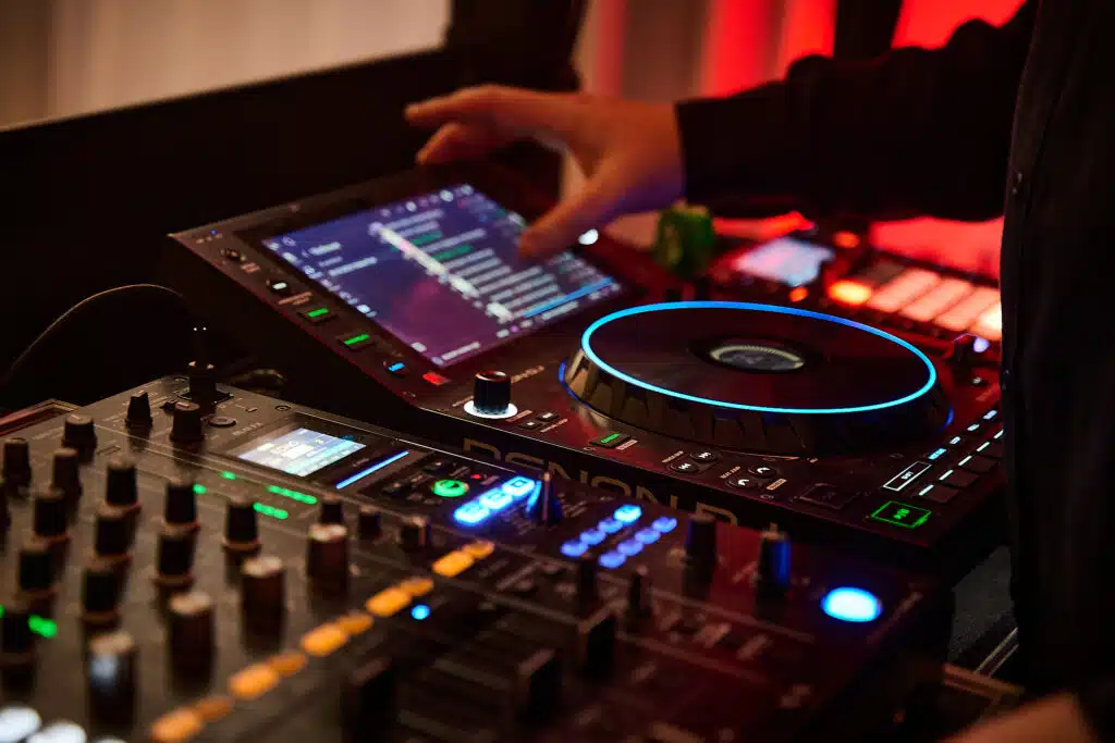 Zbliżenie na sprzęt DJ-ski podczas 18. urodzin. Widoczne są ręce DJ-a obsługujące mikser i kontroler marki Denon DJ SC5000 Prime z kolorowym ekranem dotykowym i podświetlanym jog wheelem. W tle widać fragmenty oświetlenia imprezowego w odcieniach czerwieni.