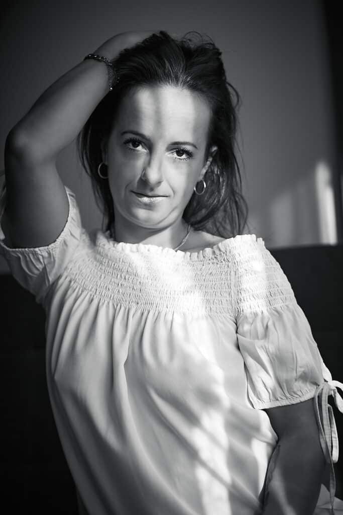 Czarno-biały portret kobiety o imieniu Justyna. Kobieta z ciemnymi włosami, ubrana w bluzkę z odkrytymi ramionami, spogląda w obiektyw. Jedną rękę ma uniesioną za głowę, drugą opiera na udzie. Na jej twarzy widoczne są cienie. W tle widać jasną ścianę i fragment ciemnej sofy.