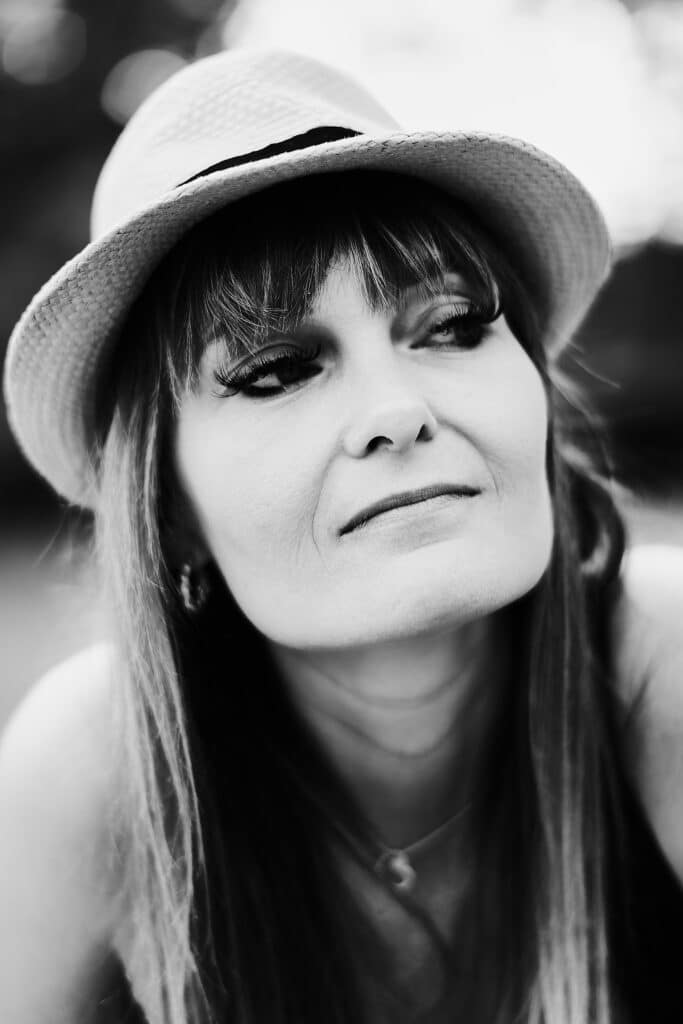 Czarno-biały portret kobiety o imieniu Kamila. Kobieta ma długie włosy z grzywką i nosi kapelusz typu fedora z ciemnym paskiem. Spogląda w górę z lekkim uśmiechem. Widoczny jest fragment jej naszyjnika i kolczyka. Tło jest nieostre.