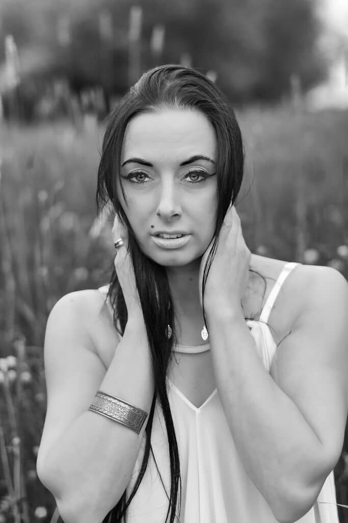 Czarno-biały portret młodej kobiety z długimi, ciemnymi włosami. Modelka patrzy prosto w obiektyw, trzymając dłonie przy szyi. Ma na sobie jasną sukienkę na ramiączkach i szeroką bransoletkę na jednym nadgarstku. Tło jest nieostre, przedstawiające łąkę z drobnymi kwiatami, co skupia uwagę na twarzy i dekolcie modelki.