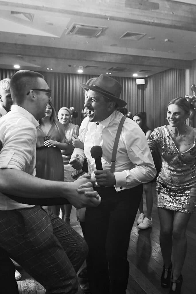 Czarno-białe zdjęcie przedstawiające wodzireja w kapeluszu i z mikrofonem w ręku, prowadzącego zabawę na 18-tych urodzinach. Wodzirej śmieje się i gestykuluje, trzymając za ręce jednego z uczestników zabawy. Wokół nich tańczą i śmieją się inni goście, w tym młode kobiety w sukienkach. W tle widać oświetlenie sali i elementy dekoracji. Zdjęcie wykonał Fotograf z Pabianic KMOPhoto.