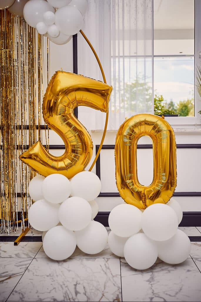 Złote balony foliowe uformowane w liczbę '50' stanowią centralny punkt dekoracji urodzinowej. Każda cyfra umieszczona jest na podstawie z białych, okrągłych balonów. Za balonami widoczne są złote, błyszczące frędzle, białe zasłony i okno w tle. Całość ustawiona jest na marmurowej podłodze.