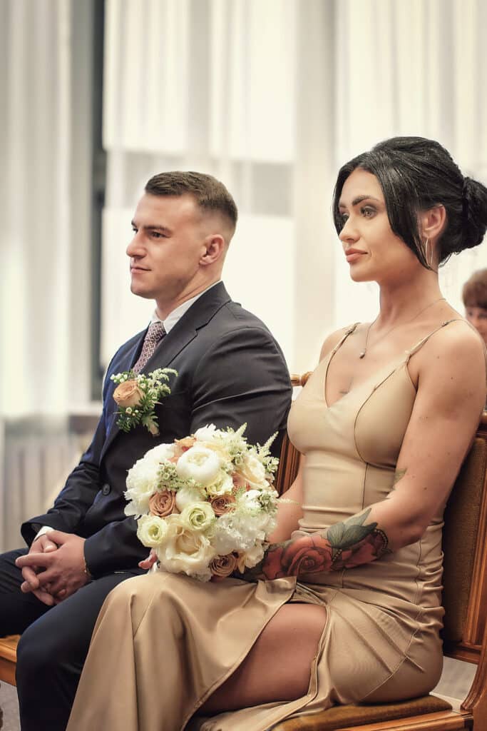 Para młoda siedzi obok siebie podczas ceremonii ślubu cywilnego. Pan młody w ciemnym garniturze z butonierką spogląda w lewo. Panna młoda w eleganckiej, satynowej sukience w kolorze szampana trzyma na kolanach bukiet ślubny z białych i herbacianych róż oraz białych piwonii i spogląda w prawo. Oboje siedzą na drewnianych krzesłach. W tle widać jasne zasłony i fragmenty innych osób