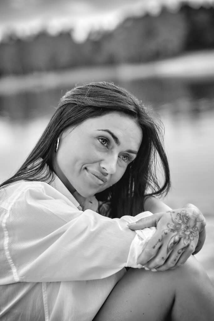 Czarno-biały portret kobiety w białej koszuli siedzącej na brzegu jeziora. Brunetka z długimi włosami spogląda w bok z delikatnym uśmiechem. Jej ręce spoczywają na kolanach, a na jednej z nich widoczny jest tatuaż o motywie roślinnym. W tle widać rozmyte jezioro i brzeg