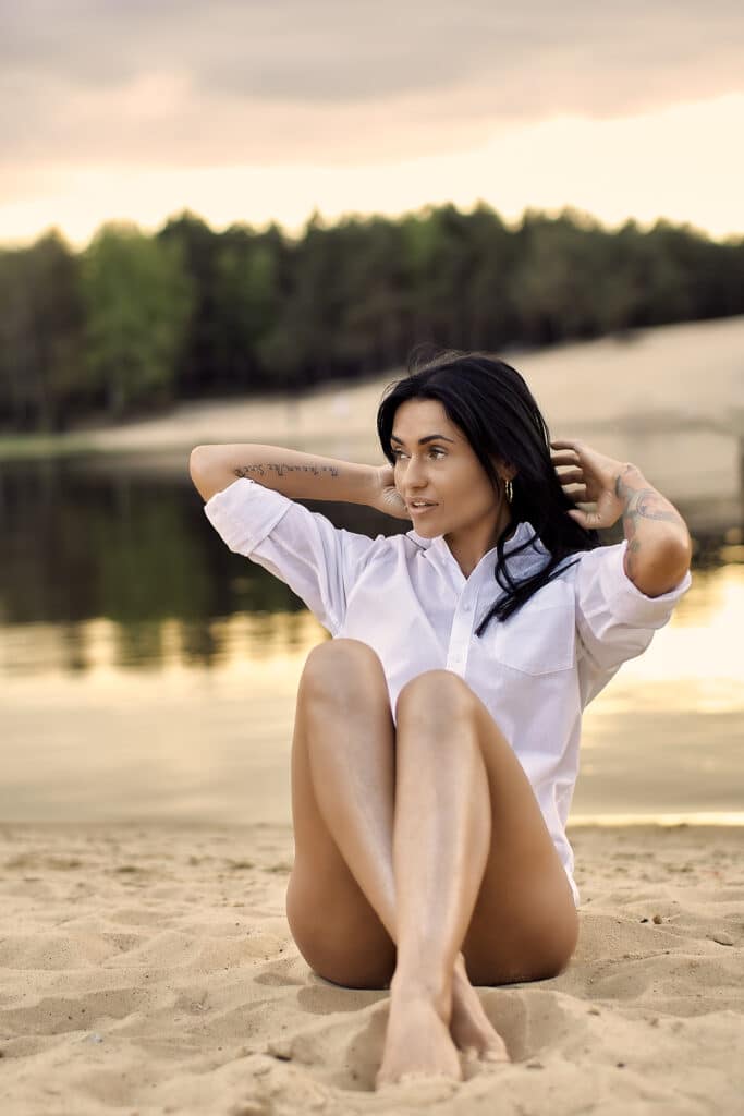 Kobieta w białej koszuli siedzi na piaszczystym brzegu jeziora z podkulonymi nogami i rękami założonymi za głowę. Brunetka spogląda w bok z lekkim uśmiechem. W tle widać spokojną wodę i zalesiony brzeg pod wieczornym niebem. Scena emanuje spokojem i naturalnością.