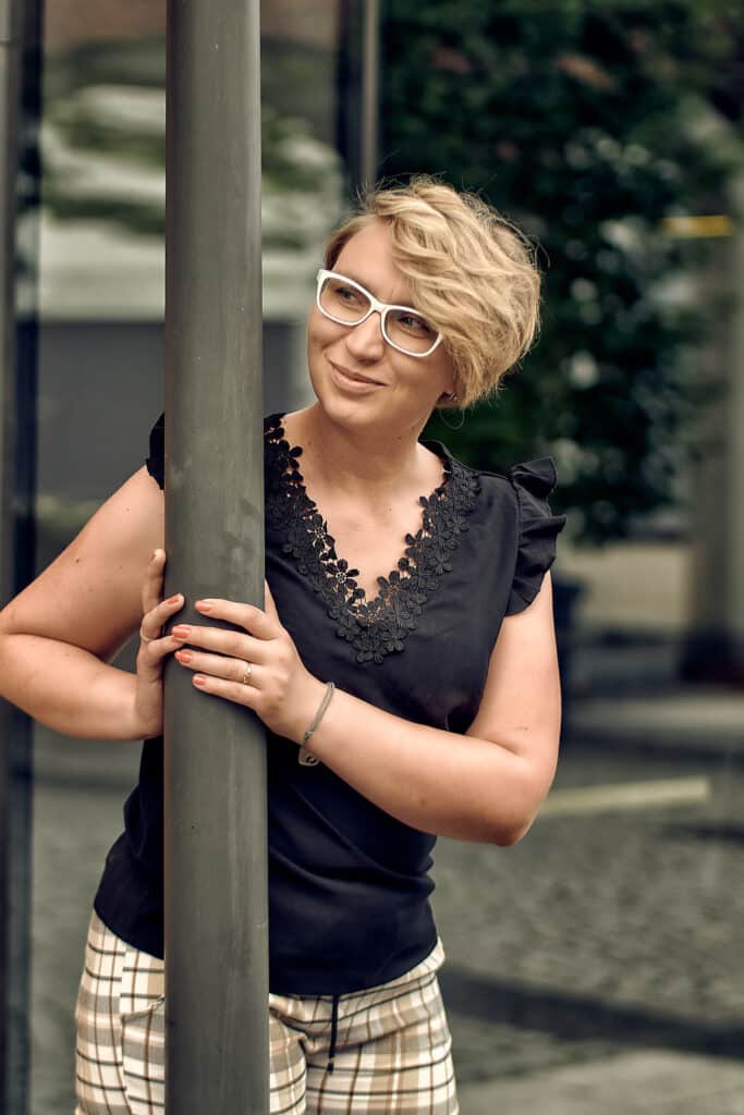 Portret biznesowy kobiety o krótkich blond włosach w białych okularach, opierającej się o szary słup. Kobieta ma na sobie czarną bluzkę z koronkowym dekoltem i wzorzyste spodnie w kratę. Spogląda w bok z lekkim uśmiechem. W tle widać rozmyty budynek z szklaną fasadą i zieleń. zdjęcie wykonał Fotograf Pabianice.
