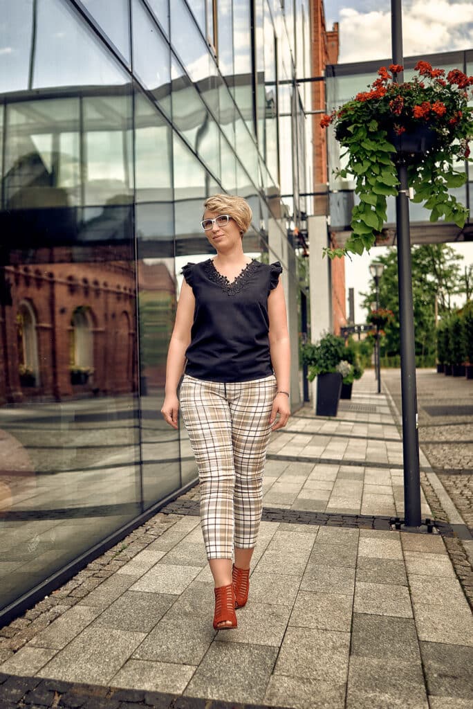 Kobieta o krótkich blond włosach i w okularach przeciwsłonecznych idzie chodnikiem wzdłuż nowoczesnego budynku z szklaną fasadą. Ma na sobie czarną bluzkę z koronkowym dekoltem i wzorzyste spodnie w kratę. Na nogach ma brązowe botki. W tle widoczny jest fragment ceglanego budynku i kwietniki z czerwonymi kwiatami. Scena jest dynamiczna i miejska.