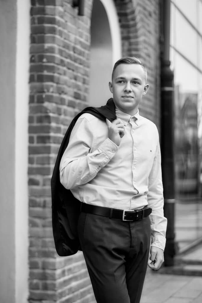 Czarno-białe zdjęcie przedstawiające młodego mężczyznę w jasnej koszuli i ciemnych spodniach z paskiem. Mężczyzna ma krótkie włosy i patrzy w stronę obiektywu. Na ramieniu ma przewieszoną ciemną marynarkę. W tle widać ceglany mur z łukowym przejściem i fragment nowoczesnego budynku z dużymi oknami
