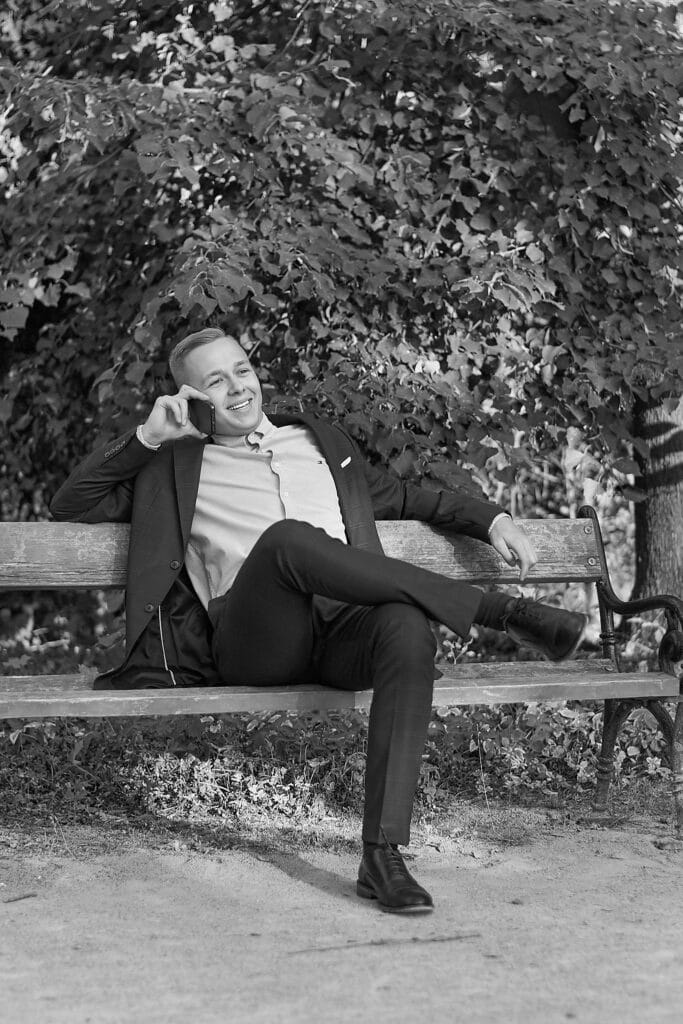 Czarno-białe zdjęcie przedstawiające mężczyznę w garniturze siedzącego na ławce w parku i rozmawiającego przez telefon. Mężczyzna jest uśmiechnięty i patrzy w górę. Ma założoną marynarkę, jasną koszulę i ciemne spodnie. Jedna noga jest założona na drugą. Za nim widać liściaste drzewo, a pod ławką trawę i ścieżkę