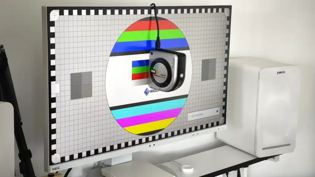 Monitor NEC z białymi ramkami wyświetla planszę testową EIZO, używaną do kalibracji monitora i oceny jakości obrazu. Na środku ekranu, w okręgu z pasami kolorów, wisi kalibrator Calibrite ColorChecker Studio, aktywnie mierzący parametry wyświetlacza. Tło planszy stanowi siatka i skala szarości, co jest kluczowe dla precyzyjnego zarządzania kolorem i zapewnienia dokładnego odwzorowania barw.