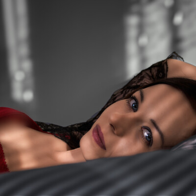 Apartamentowa sesja sensualna - Zbliżenie na twarz Moniki leżącej na ciemnej pościeli. Kobieta z jasnymi oczami i ciemnymi włosami spogląda w górę z delikatnym wyrazem twarzy. Widoczny jest fragment jej czerwonej bielizny i koronkowej narzutki. Tło jest ciemne i rozmyte, co podkreśla intymność i zmysłowość ujęcia.