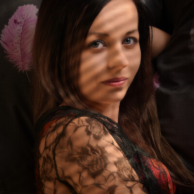 Portret Moniki w klimacie sensual. Młoda kobieta z długimi, ciemnymi włosami i jasnymi oczami spogląda w obiektyw. Na jej twarzy widoczne są cienie, prawdopodobnie od żaluzji. Ubrana jest w koronkową bluzkę. W tle widać ciemną tkaninę i delikatne, różowe pióro na poduszce, co tworzy intymną i zmysłową atmosferę.