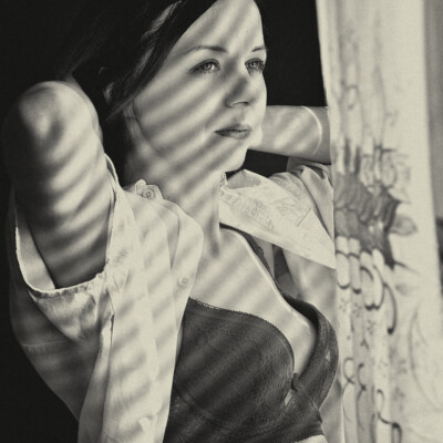 Czarno-biały portret młodej kobiety w biustonoszu i rozpiętej koszuli, stojącej przy oknie. Na jej twarzy i ciele widoczne są wyraźne cienie od żaluzji. Kobieta ma ciemne włosy i jasne oczy, a jej ręce są uniesione za głowę. Na jej boku widoczny jest tatuaż. Tło jest ciemne z wyjątkiem jasnego okna z delikatną firanką, co tworzy kontrastowe i intymne ujęcie.