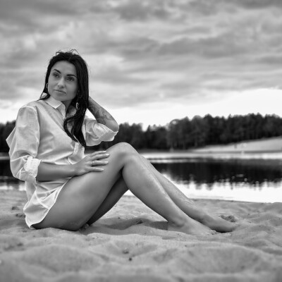 Czarno-białe zdjęcie kobiety w białej koszuli siedzącej na piaszczystej plaży. Brunetka ma podkulone nogi i jedną rękę opartą na piasku, a drugą dotyka włosów. Spogląda w bok. W tle widać spokojną wodę i zalesiony brzeg pod zachmurzonym niebem, tworząc spokojną i zamyśloną atmosferę