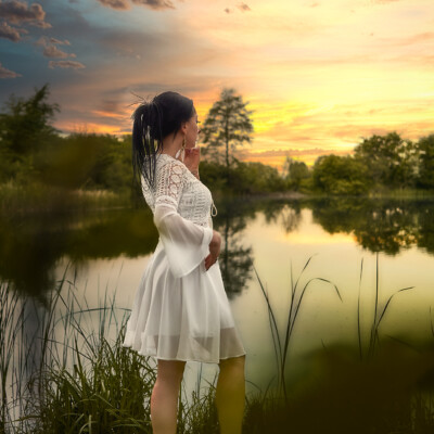 Kobieta w białej, koronkowej sukience stoi boso na brzegu jeziora o zachodzie słońca. Brunetka z włosami spiętymi w kucyk patrzy w dal, opierając dłoń o policzek. W tle widać spokojną wodę odbijającą niebo w odcieniach pomarańczy, żółci i szarości oraz zalesiony brzeg. Na pierwszym planie widoczne są trawy, co dodaje naturalności i spokoju scenie.