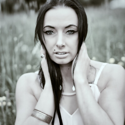 Czarno-biały portret młodej kobiety z długimi, ciemnymi włosami. Modelka patrzy prosto w obiektyw, trzymając dłonie przy szyi. Ma na sobie jasną sukienkę na ramiączkach i szeroką bransoletkę na jednym nadgarstku. Tło jest nieostre, przedstawiające łąkę z drobnymi kwiatami, co skupia uwagę na twarzy i dekolcie modelki.