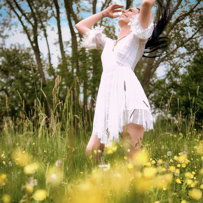 Kobieta w białej, koronkowej sukience stoi na łące pełnej żółtych kwiatów. Brunetka z uniesionymi rękami i rozpuszczonymi włosami spogląda w górę z uśmiechem. W tle widać drzewa i błękitne niebo z chmurami. Scena emanuje radością, swobodą i naturalnym pięknem.