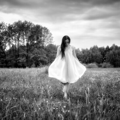 Modelka Marika - Czarno-biała fotografia kobiety w jasnej, zwiewnej sukience idącej przez łąkę. Brunetka z długimi włosami spogląda w dół, trzymając sukienkę za boki. Jest boso. W tle widać ciemne drzewa i pochmurne niebo, co nadaje zdjęciu melancholijny i eteryczny nastrój.
