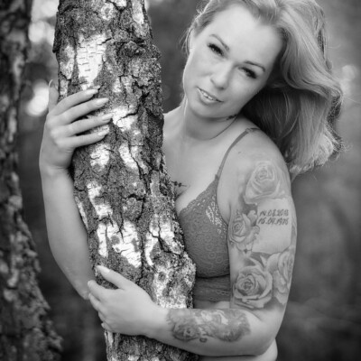 Czarno-biały portret modelki Pauliny w bieliźnie, pozującej przy drzewie. Kobieta z długimi, falującymi włosami obejmuje drzewo, spoglądając w obiektyw. Na jej ramieniu widoczne są tatuaże. Tło jest rozmyte, co skupia uwagę na modelce.