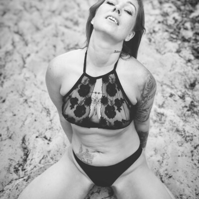 Czarno-białe zdjęcie modelki w delikatnej bieliźnie, pozującej na piasku. Kobieta klęczy z głową odchyloną do tyłu i przymkniętymi oczami. Jej ciało jest skierowane w stronę obiektywu. Na jej skórze widoczne są tatuaże. Tło jest piaszczyste i nieostre, co podkreśla postać modelki.