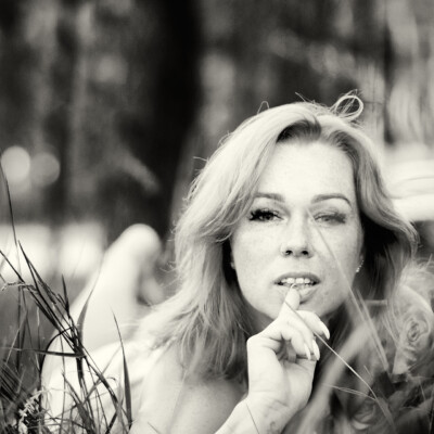 Czarno-biały portret kobiety z długimi, falującymi włosami, leżącej w trawie. Modelka patrzy w obiektyw z palcem przy ustach. Tło jest nieostre, z widocznymi źdźbłami trawy na pierwszym planie, co tworzy intymną i naturalną atmosferę.