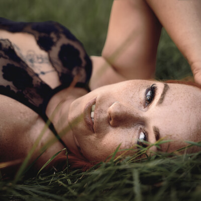 Zbliżenie na twarz rudej modelki z piegami, leżącej w trawie. Kobieta patrzy w górę z delikatnym uśmiechem. Widoczny jest fragment jej czarnej, koronkowej bielizny oraz dłoń oparta na trawie. Tło jest zielone i rozmyte, podkreślając naturalność i intymność sceny.