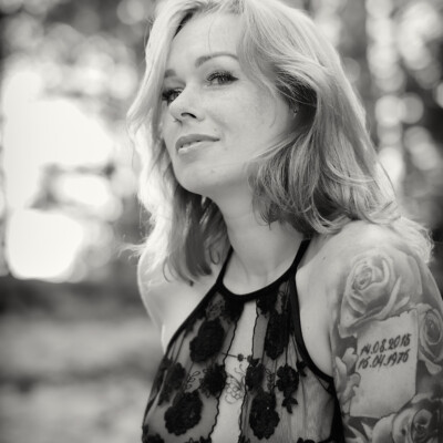 Czarno-biały portret modelki Pauliny z długimi, falującymi włosami, ubranej w delikatną, koronkową bieliznę. Kobieta spogląda w górę z subtelnym uśmiechem. Na jej ramieniu widoczny jest tatuaż z motywem róży i datami. Tło jest rozmyte, z jasnymi punktami światła, co nadaje zdjęciu miękki i intymny charakter.