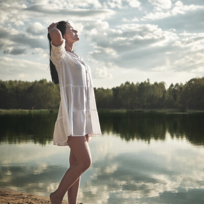 Sesja Kobieca - kobieta w białej, zwiewnej sukience stoi boso na piaszczystym brzegu jeziora. Brunetka ma ręce założone za głowę i patrzy w górę, w stronę nieba. W tle widać spokojną wodę odbijającą chmury i zalesiony brzeg. Scena jest pełna spokoju i naturalnego piękna