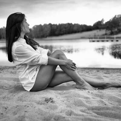 Czarno-białe zdjęcie kobiety w białej koszuli siedzącej boso na piaszczystym brzegu jeziora. Brunetka z profilu spogląda w stronę wody, obejmując nogi rękami. W tle widać spokojną powierzchnię jeziora i zalesiony brzeg pod pochmurnym niebem. Scena emanuje spokojem i zamyśleniem.