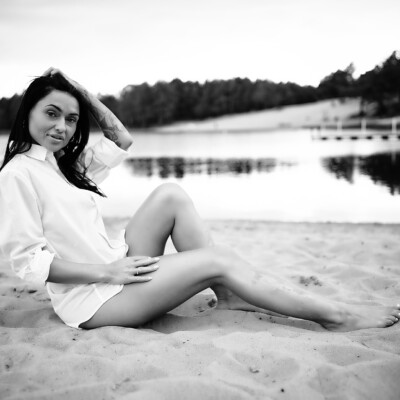 Czarno-białe zdjęcie kobiety w białej koszuli siedzącej na piaszczystym brzegu jeziora. Brunetka z jedną nogą wyprostowaną, a drugą ugiętą, opiera się jedną ręką o piasek, a drugą dotyka włosów. Spogląda w stronę obiektywu. W tle widać spokojną wodę i zalesiony brzeg pod pochmurnym niebem, tworząc swobodną i naturalną scenę