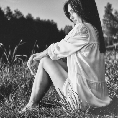 Czarno-białe zdjęcie kobiety w białej, luźnej koszuli siedzącej na trawie. Brunetka z podkulonymi nogami i rękami splecionymi na kolanach spogląda w dół. W tle widać ciemne drzewa i jasne niebo, tworząc spokojną i melancholijną scenę.