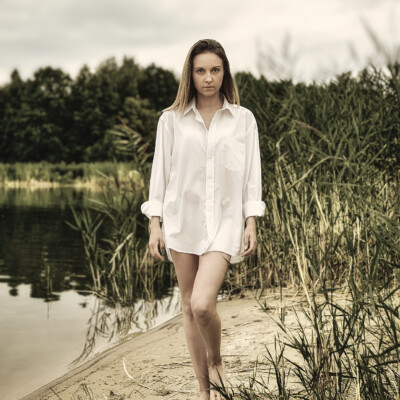 Monika - Kobieta w białej, męskiej koszuli idzie boso brzegiem jeziora. Blondynka z długimi włosami spogląda w dół. W tle widać spokojną wodę i zalesiony brzeg pod pochmurnym niebem. Na pierwszym planie widoczne są wysokie trawy, co tworzy naturalną i spokojną scenę.