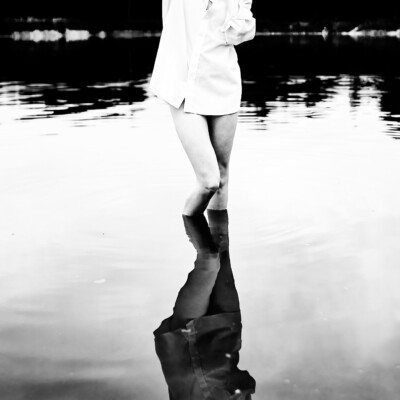 Czarno-białe zdjęcie kobiety w białej, rozpiętej koszuli stojącej w wodzie. Blondynka z długimi włosami ma jedną rękę uniesioną nad głową i spogląda w dół, na swoje odbicie w spokojnej powierzchni wody. W tle widać ciemny brzeg i pochmurne niebo, co tworzy minimalistyczną i refleksyjną scenę.