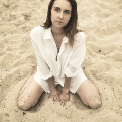 Kobieta w białej, rozpiętej koszuli klęczy na piaszczystej plaży. Blondynka z długimi włosami spogląda prosto w obiektyw z poważnym wyrazem twarzy. Jej dłonie spoczywają na piasku przed nią. Tło jest piaszczyste i lekko rozmyte, co skupia uwagę na modelce.