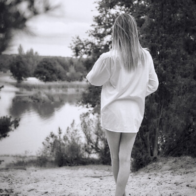 Czarno-białe zdjęcie kobiety w białej, męskiej koszuli stojącej tyłem na piaszczystym brzegu jeziora. Blondynka z długimi włosami spogląda w stronę wody. W tle widać spokojną powierzchnię jeziora i zalesiony brzeg pod pochmurnym niebem. Scena emanuje spokojem i kontemplacją.
