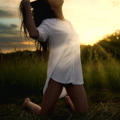 Kobieta klęczy boso na łące o zachodzie słońca. Brunetka w białej, rozpiętej koszuli ma uniesione ręce, trzymając włosy nad głową i spogląda w górę. W tle widać niebo w odcieniach pomarańczy i szarości oraz zarysy drzew. Promienie zachodzącego słońca dodają scenie ciepła i intymności.