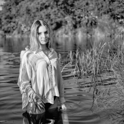 Czarno-białe zdjęcie kobiety w białej, rozpiętej koszuli stojącej w wodzie jeziora. Blondynka z długimi włosami spogląda w stronę obiektywu. Woda sięga jej do ud. W tle widać brzeg z drzewami i wysokimi trawami. Scena jest spokojna i naturalna.