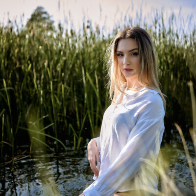 Kobieta w białej koszuli stoi w płytkiej wodzie wśród wysokich traw. Blondynka z długimi włosami spogląda w stronę obiektywu. Woda odbija niebo i otaczającą roślinność. Scena jest naturalna, spokojna i pełna letniego światła.