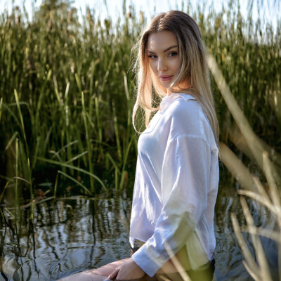 Martyna - Kobieta w białej koszuli stoi w płytkiej wodzie porośniętej wysokimi trawami. Blondynka z długimi włosami odwraca się i spogląda przez ramię w stronę obiektywu. Jej dłoń zanurzona jest w wodzie. W tle widać błękitne niebo i zieleń. Scena jest naturalna, spokojna i pełna letniego światła.
